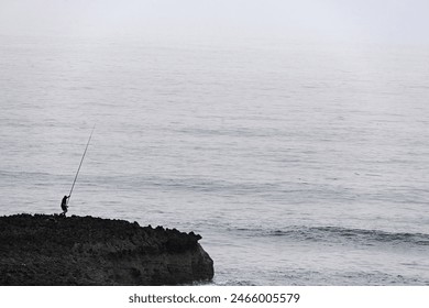 A solitary fisherman with a fishing rod stands on a rocky coastline, casting into the misty sea. The tranquil and serene setting highlights the quiet pursuit of fishing against the vast, calm ocean. - Powered by Shutterstock