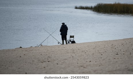 A solitary fisherman stands near the water's edge, casting a line into the tranquil lake under a clear sky. The sandy bank surrounds him - Powered by Shutterstock