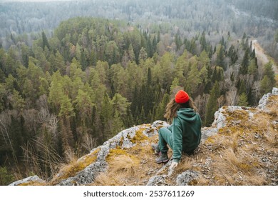 A solitary hiker in a green hoodie and red hat sits on a rocky ledge, lost in contemplation, admiring the lush forest and meandering path below, surrounded by nature - Powered by Shutterstock