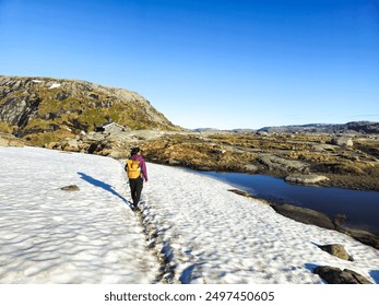 A solitary hiker traverses a snowy path, framed by lush hills and a tranquil lake under a bright blue sky. Lysefjord Kjeragbolten Kjerag Norway - Powered by Shutterstock