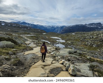 A solitary hiker treks along a rocky path, surrounded by Norway's stunning mountains and serene lakes under a cloudy sky. Trolltunga, Norway - Powered by Shutterstock