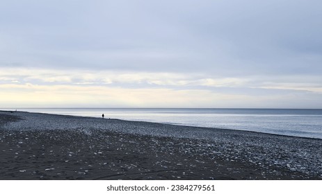 A solitary individual is captured in the distance, traversing the desolate, rocky beach that meets the cold blue embrace of the sea, creating a poignant scene of isolation and the vastness of nature