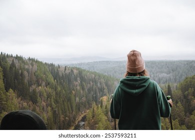 A solitary individual in a cozy hoodie and stylish beanie stands on a cliff, lost in thought while gazing at the lush green forest beneath a dramatic cloudy sky - Powered by Shutterstock