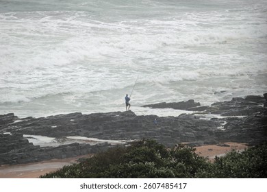 A solitary individual fishing with a rod on a rocky seashore while waves crash nearby under a cloudy sky, evoking a sense of serenity and connection with nature.