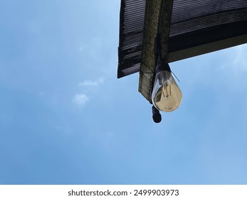 A solitary light bulb hangs from a weathered roof, casting a dramatic silhouette against a vibrant blue sky. light bulb isolated on a blue sky - Powered by Shutterstock