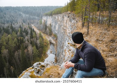 A solitary man finds himself perched at the very edge of a steep, rocky cliff, deeply gazing at the serene river winding below, all while being surrounded by a breathtaking natural landscape - Powered by Shutterstock