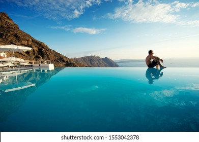 Solitary man sitting on the edge of an infinity pool looking out over the scenic Mediterranean view of the Santorini caldera, Greece