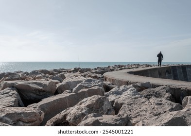 A solitary person walks along a rocky pier, overlooking a calm sea under a vast, clear sky. The scene feels peaceful and introspective, with rugged rocks contrasting the serene horizon. - Powered by Shutterstock