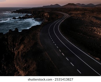 A solitary road winds along the rugged coastline during dusk, framed by dramatic cliffs and the ocean's waves crashing against the rocks.  - Powered by Shutterstock