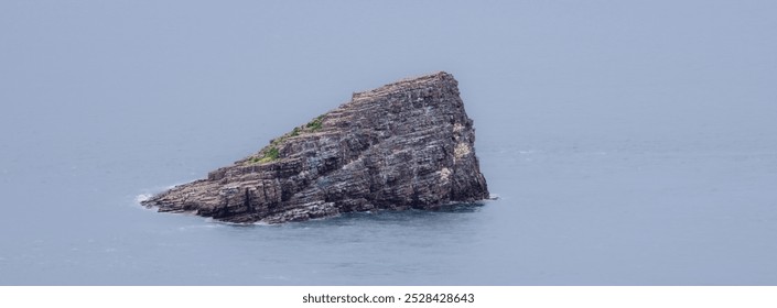 A solitary rocky island in Brittany rising from calm ocean waters, surrounded by gentle waves. The island features rugged cliffs and sparse greenery on top, set against a gray sky. - Powered by Shutterstock