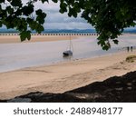 A solitary sailboat anchored on a serene, sandy shore at Arnside Cumbria, under a canopy of lush green leaves on a cloudy day.