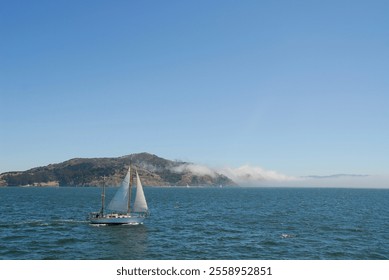 A solitary sailboat drifts through the foggy San Francisco Bay,its sails barely catching the light breeze, while the dense mist completely obscures the Golden Gate, leaving only a blank, muted horizon - Powered by Shutterstock