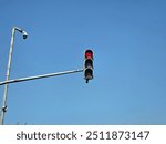 A solitary traffic light showing a red signal against a clear blue sky. The traffic light is mounted on a metal pole, and a security camera is visible nearby, highlighting urban infrastructure.