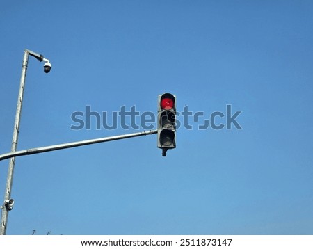 A solitary traffic light showing a red signal against a clear blue sky. The traffic light is mounted on a metal pole, and a security camera is visible nearby, highlighting urban infrastructure.