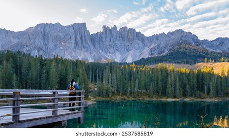 A solitary traveler stands on a wooden bridge, gazing at the serene turquoise waters reflecting the majestic Dolomites. Lake Carezza or Karersee Dolomites in Italy. - Powered by Shutterstock