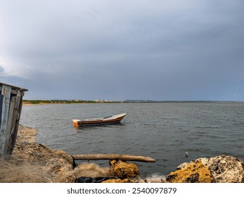 A solitary wooden boat floats on the calm waters of a river, set against a cloudy sky and a rugged shoreline. The image captures the quiet atmosphere of an overcast day by the river, with distant gree - Powered by Shutterstock