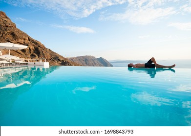 Solitary young man resting on the edge of an infinity pool overlooking a scenic Mediterranean view of the Santorini caldera, Greece