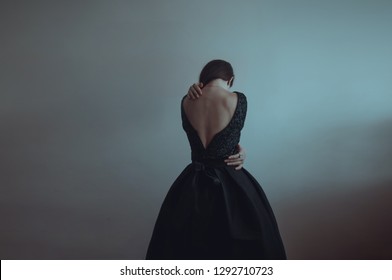 Solitude. Lonely Girl Stands Near The Gray Wall. Loneliness, Sadness, Depression, Disappointment. Woman In Vintage Black Dress. Girl Is Afraid And Hugs Herself. Dramatic Picture. Pain.