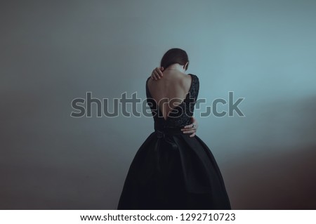 Solitude. Lonely girl stands near the gray wall. Loneliness, sadness, depression, disappointment. Woman in vintage black dress. Girl is afraid and hugs herself. Dramatic picture. Pain.