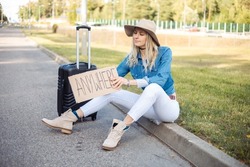 Solitude, Upset, Melancholy Blond Woman In Hat Hitchhiking, Sit On Curb By Road, Holding Carton Board Anywhere With Suitcase Luggage, Car Waiting. Adventure And Travelling On Holiday Vacation Alone. 