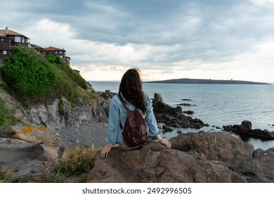 Solo Traveler with Black Hair Sitting on Rocky Cliff Overlooking Seascape and Seaside Village in Bulgaria, Enjoying a Relaxing Vacation by the Coastline with Backpack, Jeans, and a Sense of Freedom - Powered by Shutterstock