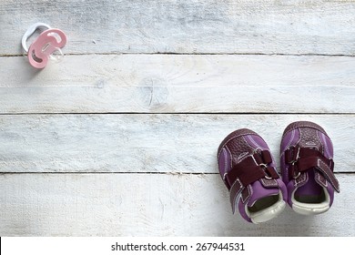 Some Baby Stuff On A White Wood, Top View. A A Pacifier And Lilac Shoes.