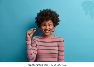 Some more, please. Cheerful smiling African American woman gestures small size with fingers, asks for little bit time, measures too small object, shows something minimum dressed casually stands indoor