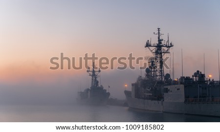 Some navy frigates and destroyers in port during a cold and foggy morning in a naval base. A fog bank between ships in port during a cold sunrise.