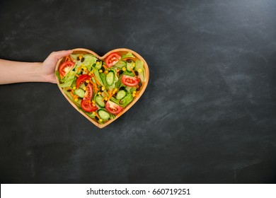 Somebody Hand Showing Nutrition Vegetable Healthy Food In Wooden Heart Shaped Plate On Black Chalkboard With High Angle View Photo.