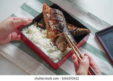 Someone Hand Using Chopsticks Trying To Pick A Grilled Saba Or Mackerel Fish Served With Cooked Rice In Square Bento Box On White And Green Striped Placemat On White Table