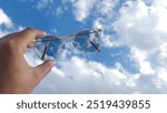 Someone is holding round-framed glasses with photochromic lenses, against a backdrop of a bright, cloudy sky.