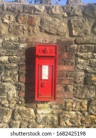 Somerset, England, UK - 24th March 2020: Sunshine Falls On A Traditional Red Letter Box Set Into A Tall Stone Wall. The Royal Cypher Initials G. R. Stand For George Rex (Rex Meaning ‘king’ In Latin).