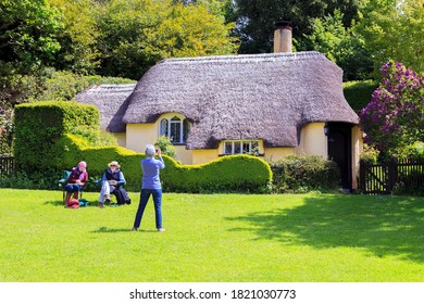 Somerset, Uk, England May 20, 2015:  Female Tourist Photographer Taking A Photograph Of A Thatched Roof Cottage In A Typical English Village  Elderly Couple Sitting Down Sketching The Scenic Views 