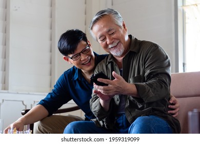 Son Cuddle Dad While Using Mobile Phone, Smiling And Laughing Together. Old Asian Man Learning To Use Smartphone By Young Asian Man Suggest Him. Father's Day And Happy Family Concept.