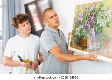 Son And Dad Hanging Picture Of Lilac On Wall. Son Holding Hammer In Hands.