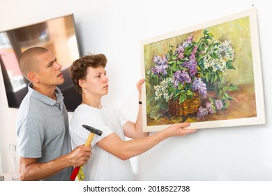 Son And Dad Hanging Picture Of Lilac On Wall. Son Holding Picture In Hands.