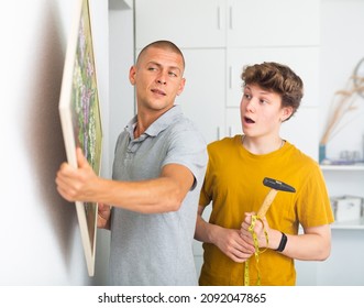 Son And Dad Hanging Picture Of Lilac On Wall. Son Holding Hammer In Hands.