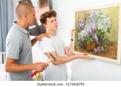 Son And Dad Hanging Picture Of Lilac On Wall. Son Holding Picture In Hands.