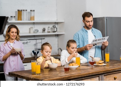 Son And Daughter Eating Oatmeal While Father Reading Travel Newspaper In Kitchen