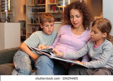 Son and daughter with their mother sit on sofa and read book in room; focus on woman