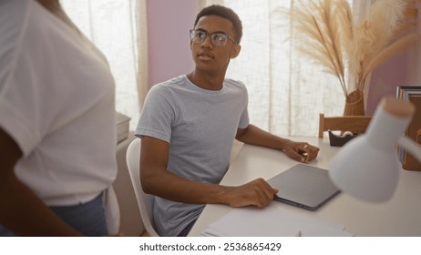 Son in glasses looks up at his mother while sitting at a desk in a cozy, sunlit living room, highlighting their close african american family bond and indoor home interaction. - Powered by Shutterstock