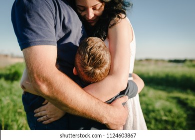 The Son Hugging Parents On Nature. Mom, Dad And Boy Walk In The Grass. Happy Young Family Spending Time Together, Outside, On Vacation, Outdoors. The Concept Of Family Holiday.