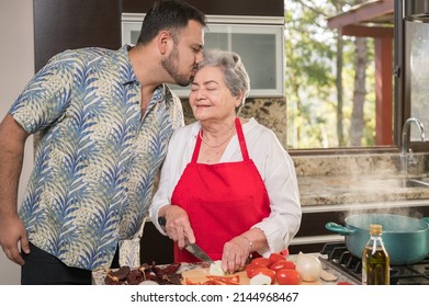 A Son Kisses His Mother On The Forehead Showing His Love, While She Was Preparing Food In Her Kitchen.