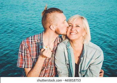 Son Kisses, Hugs His Mom In Summer Day, Outdoors. Mother And Her Adult Child Having Fun Together. Mothers Day Concept. Happiness Family Sitting On Blue Water Background. Love Concept, Emotion.