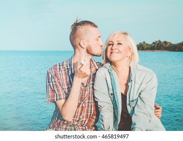 Son Kisses, Hugs His Mom In Summer Day, Outdoors. Mother And Her Adult Child Having Fun Together. Mothers Day Concept. Happiness Family Sitting On Blue Water Background. Love Concept, Emotion.