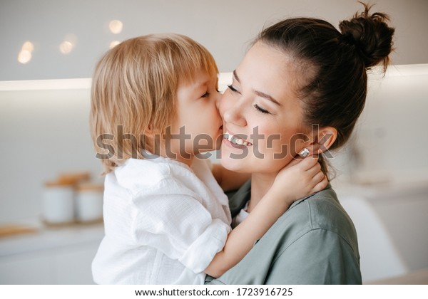 El hijo se besa en la mejilla, mamá se alegra de verla en el fondo de la cocina en casa. Niño sentado en las manos de una joven. Sonrisa en la cara de cerca.