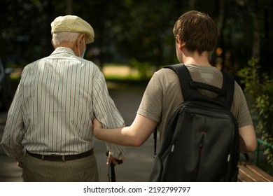 Son Leads His Father Down Street. Grandson Holds His Grandfather's Arm. Man Helps Pensioner. Old Man Is Holding On To Walking Stick. Pensioner On Street. Old Age In Russia.