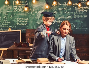 Son And Mother Have School Lesson. Genius Boy Help Woman Write Homework At Desk. Little Child In Big Suit Coat And Graduation Cap Teach Woman. Back To School. Teaching And Learning Concept.