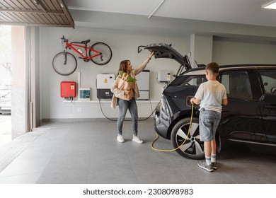Son plugs EV charger from charging station to electric vehicle in private home while mother takes groceries from the car on daytime. Sustainable Alternative Lifestyle. Horizontal copy-space.