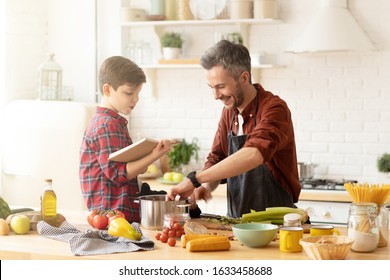 Son Sitting On Table Reading Funny Book To Dad Cooking Dinner At Modern Bright Loft Kitchen. Family Spend Time At Home. Parent And Child Communication. Good Relationships. Happy Life. Fun Recreation